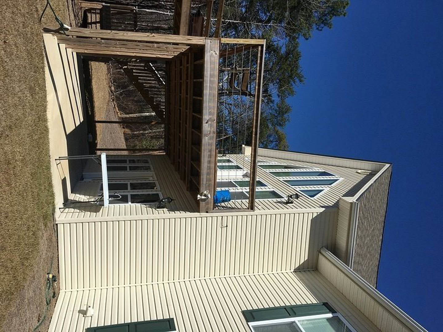 Modern two-story house with composite siding, large windows, elevated wooden deck, metal roof, and surrounding trees under clear sky