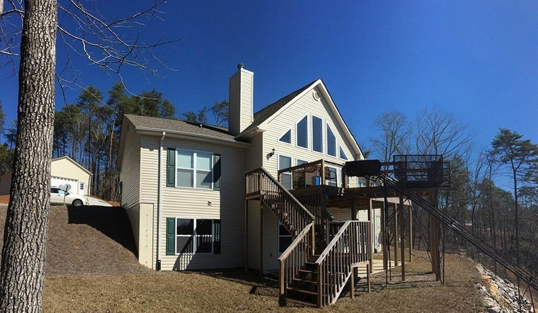 Wood siding home with elevated wooden deck, outdoor staircase, large windows, and mature trees in the yard