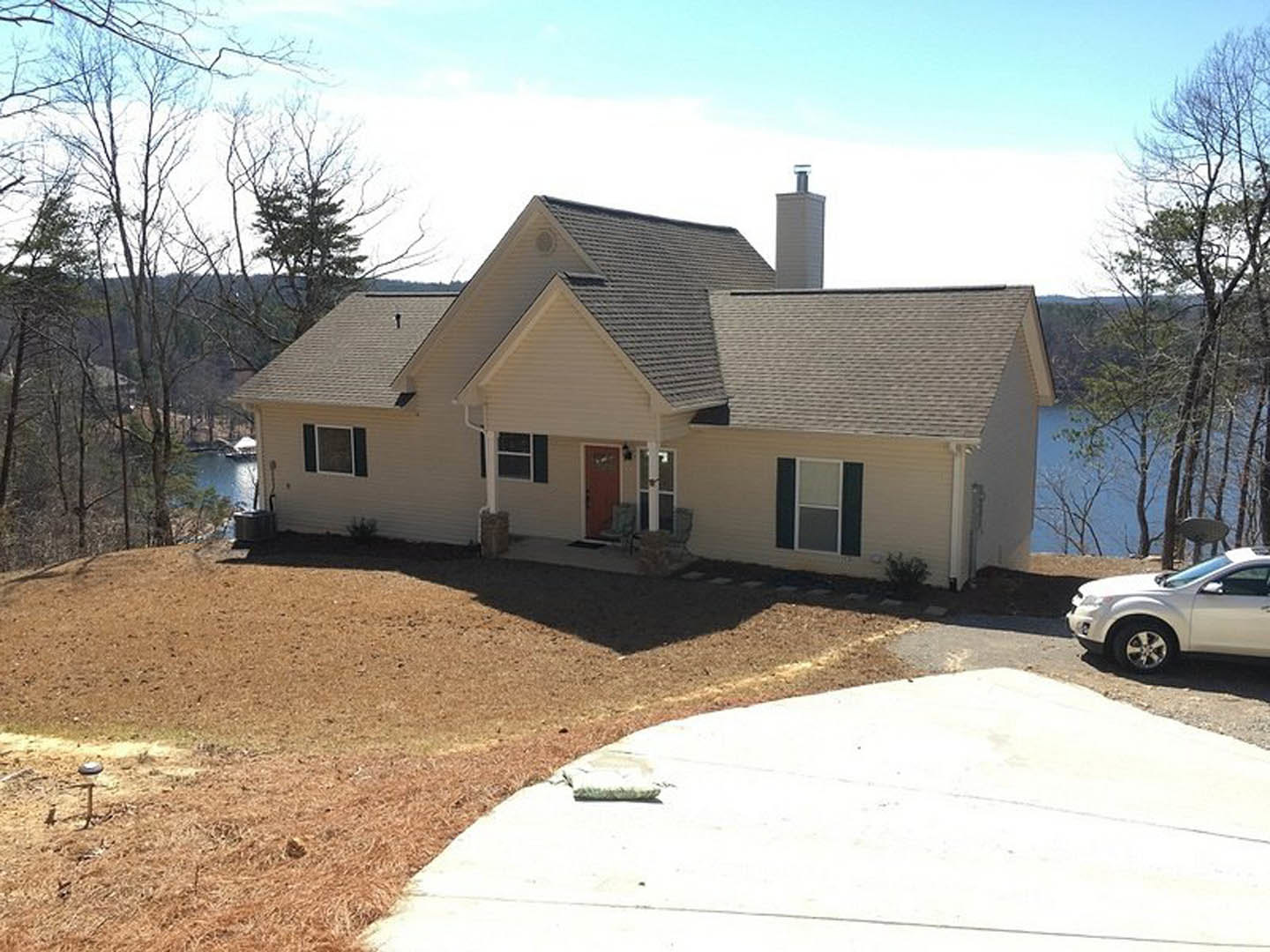 Two-story house with gray siding and brick chimney overlooking a lake, white-framed windows, white car parked on roadside, turtle on sidewalk near landscaped lawn