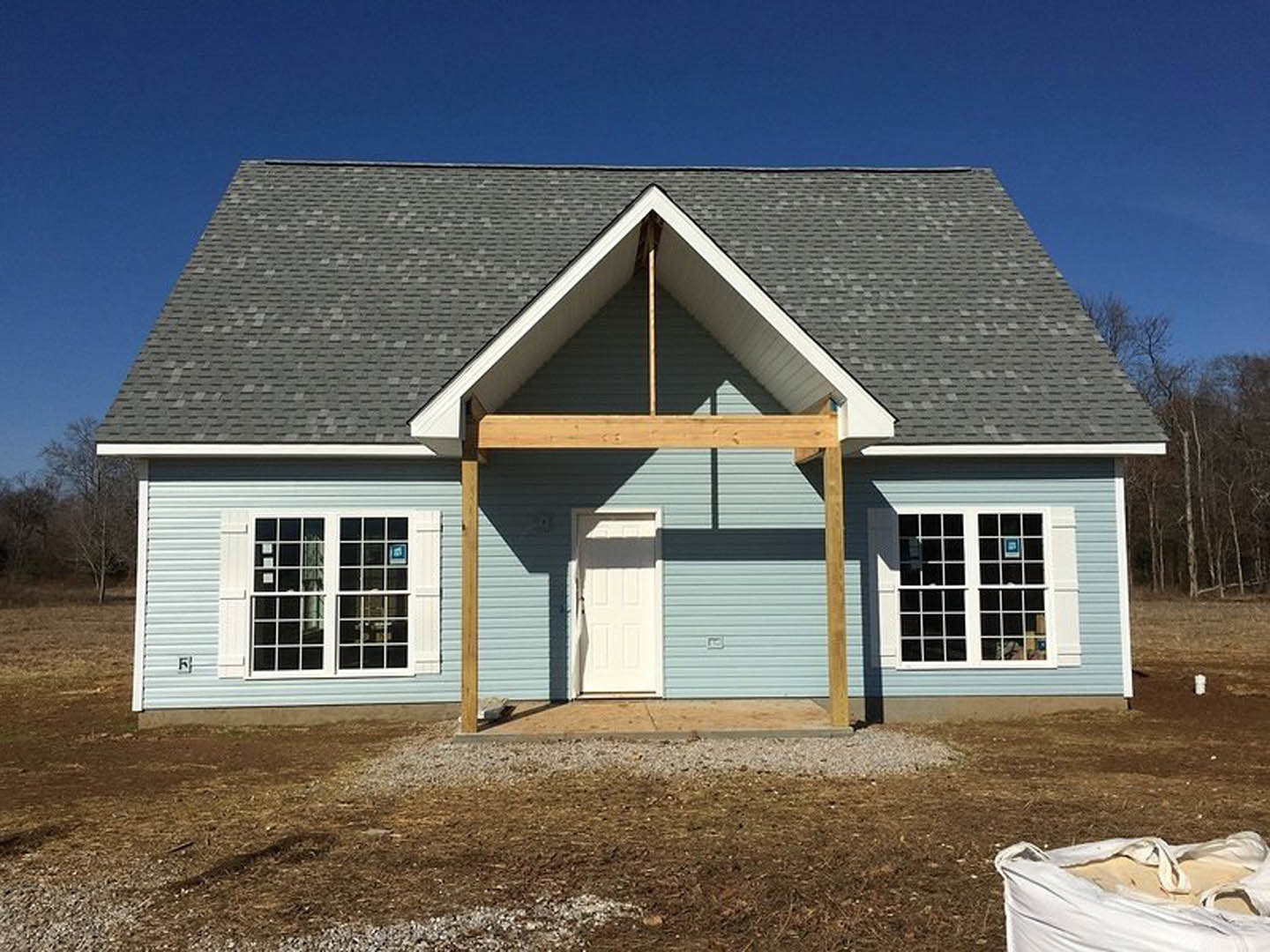 Wood-framed house under construction with exposed beams, white paneled door, multi-pane window, roof overhang, and sand bag in grassy field