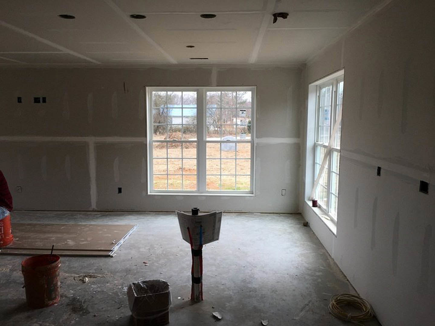 Sunlit room with plaster walls, large window overlooking green yard, light flooring, black trash can with plastic liner near wall