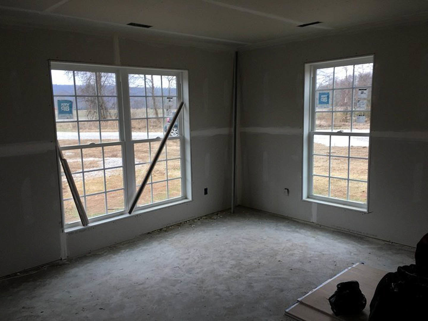 Spacious room featuring large windows, smooth concrete flooring, plaster walls, and natural daylighting; a wooden surface with a black bag and a window displaying a blue and white