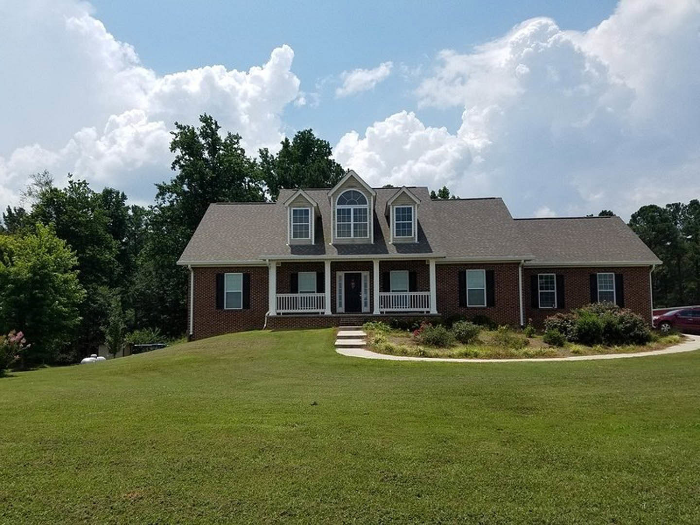 Brick exterior home with white-framed windows, covered front porch, manicured lawn, stone walkway, mature trees, cloudy sky, and a parked car partially visible.