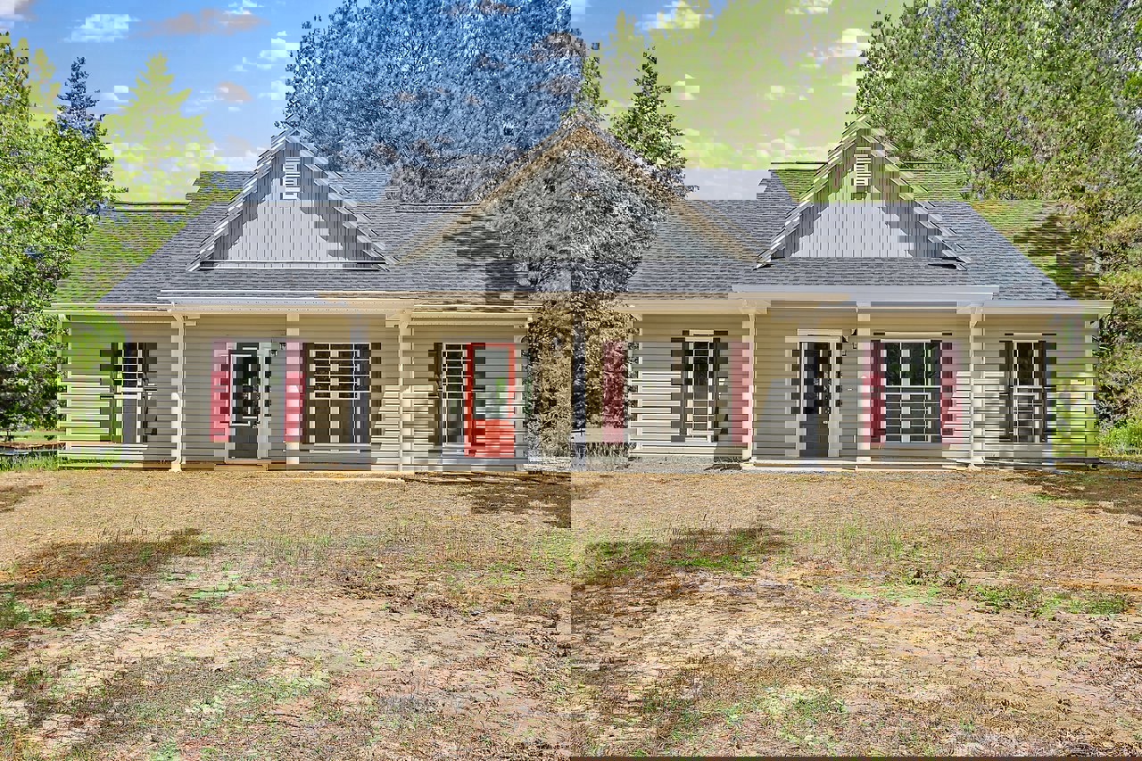 Two-story house with red doors, white columns, and white-framed windows, set on a dirt yard with a small white cottage visible in the background.