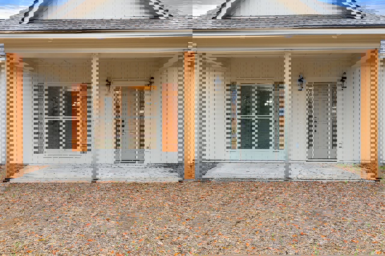 Front porch of a custom home featuring grey door, white siding, glass door with light fixture, black door handle, wood panel detail, and scattered leaves on the ground