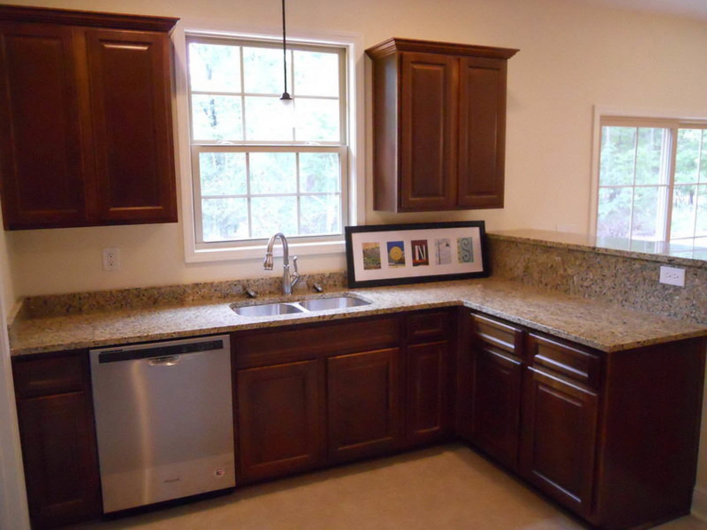Granite countertops with undermount sink and chrome faucet, stainless steel dishwasher with black handle, white cabinetry, framed picture on wall, window letting in natural light