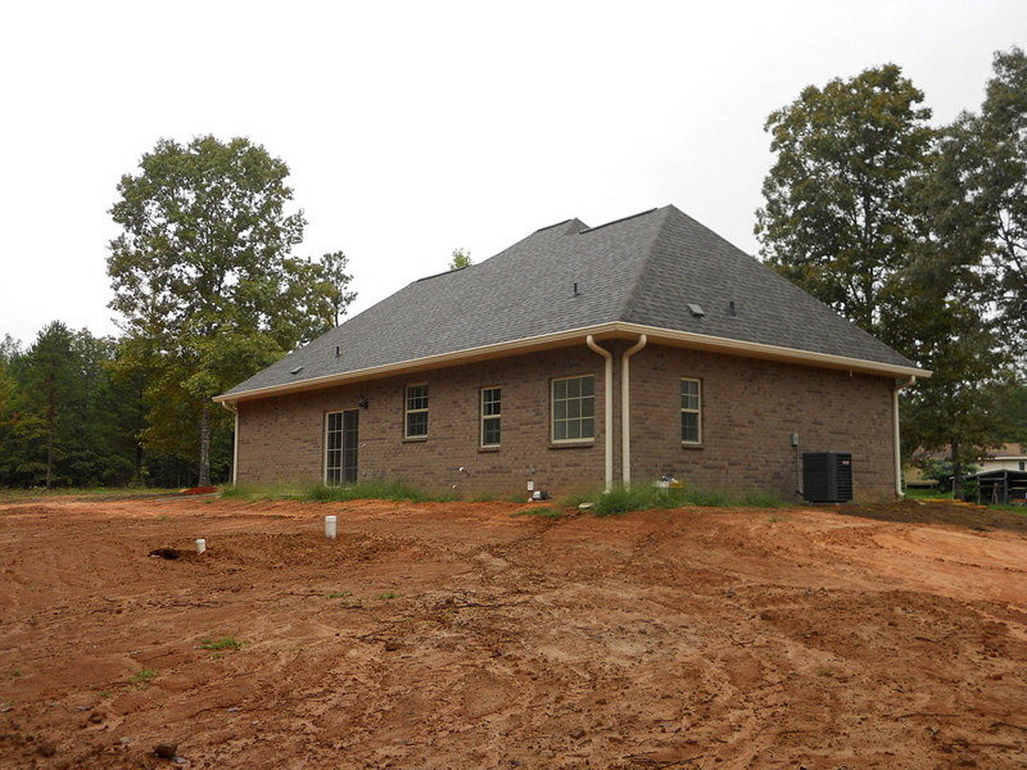 Partially built brick house with shingle roof, row of windows, dirt yard with patches of grass, black electrical box, and leafy tree in front