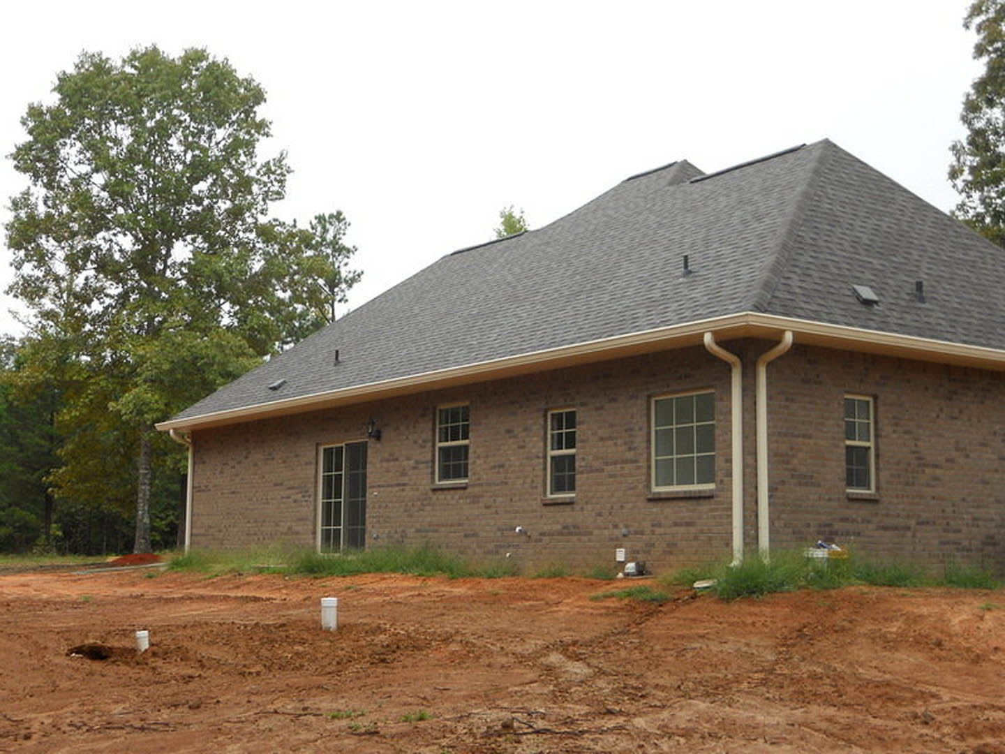 Brick house under construction with white-framed multi-pane windows, gutter system, dirt lot in foreground, and mature trees surrounding exterior