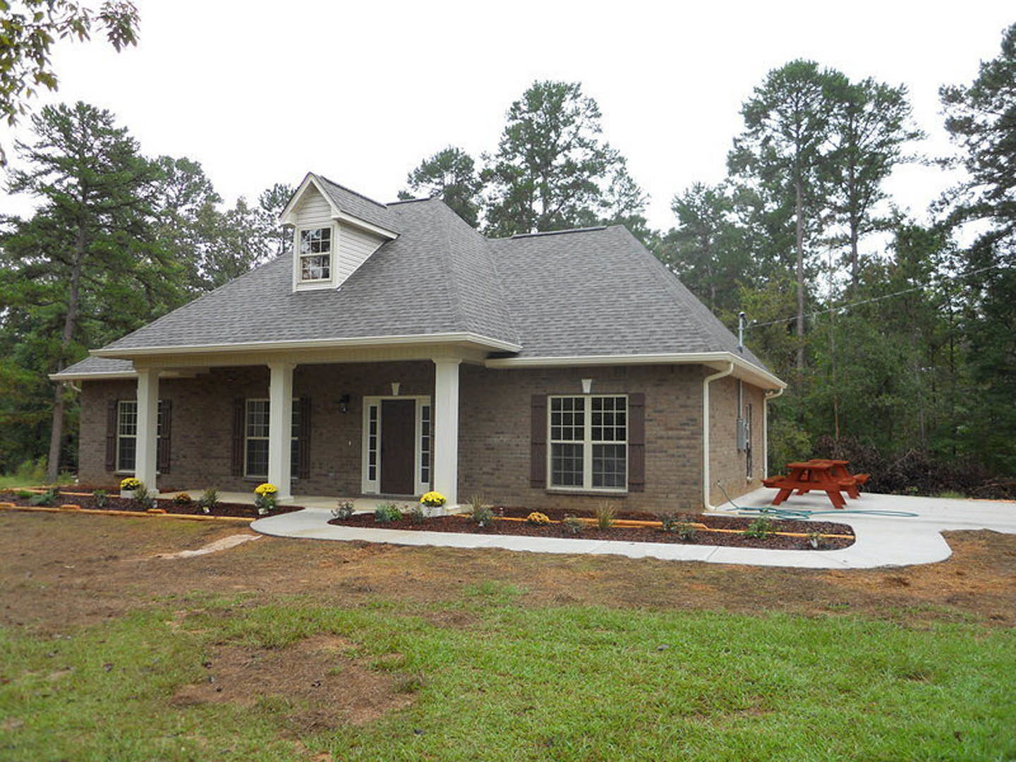 Red picnic table with white stripes on a green lawn in front of a cottage-style house featuring a porch, white-trimmed door, and multi-pane window.