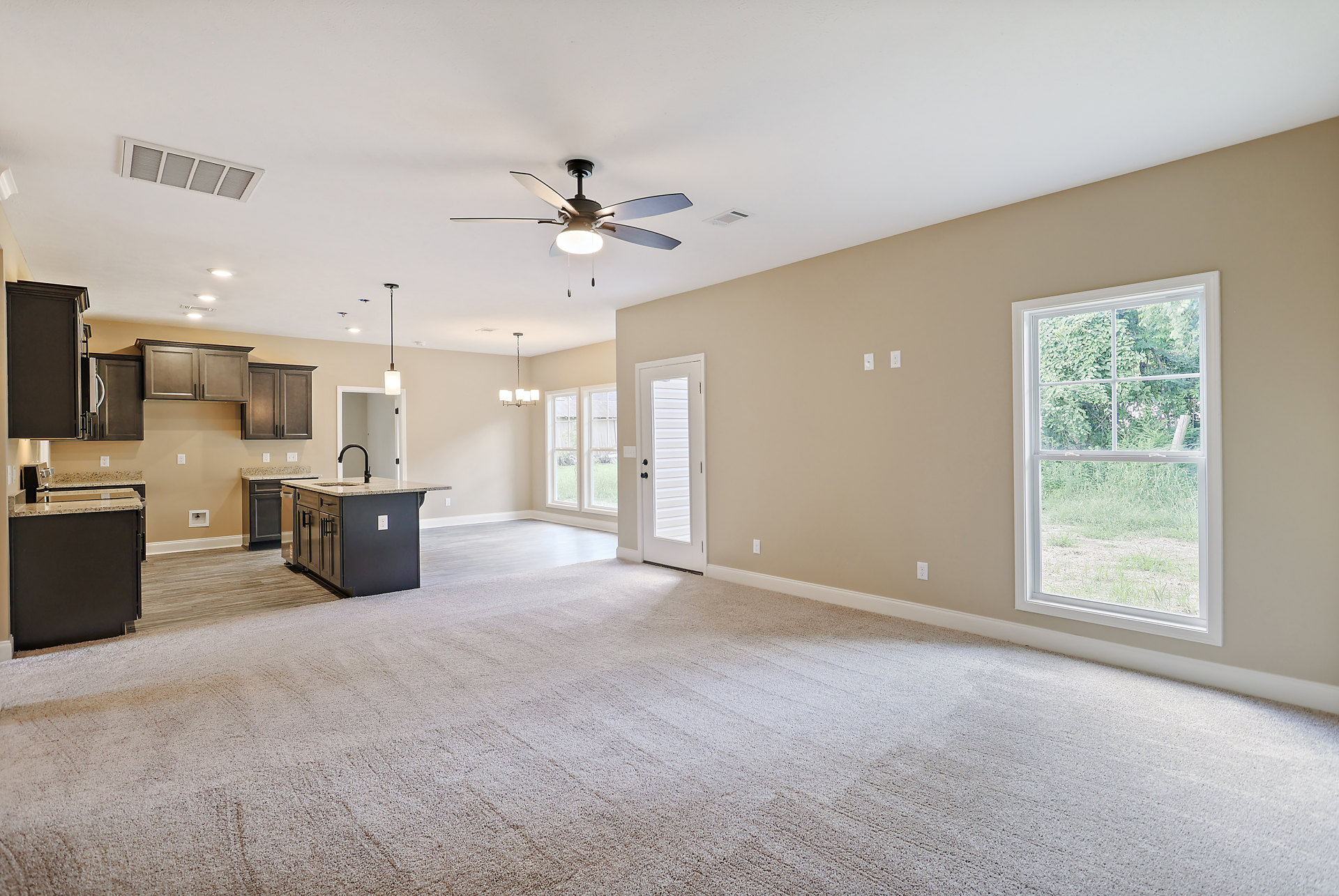 Open living room with adjacent kitchen, ceiling fan with light, window overlooking trees and grass, white door with black knobs, kitchen island featuring sink, black and white