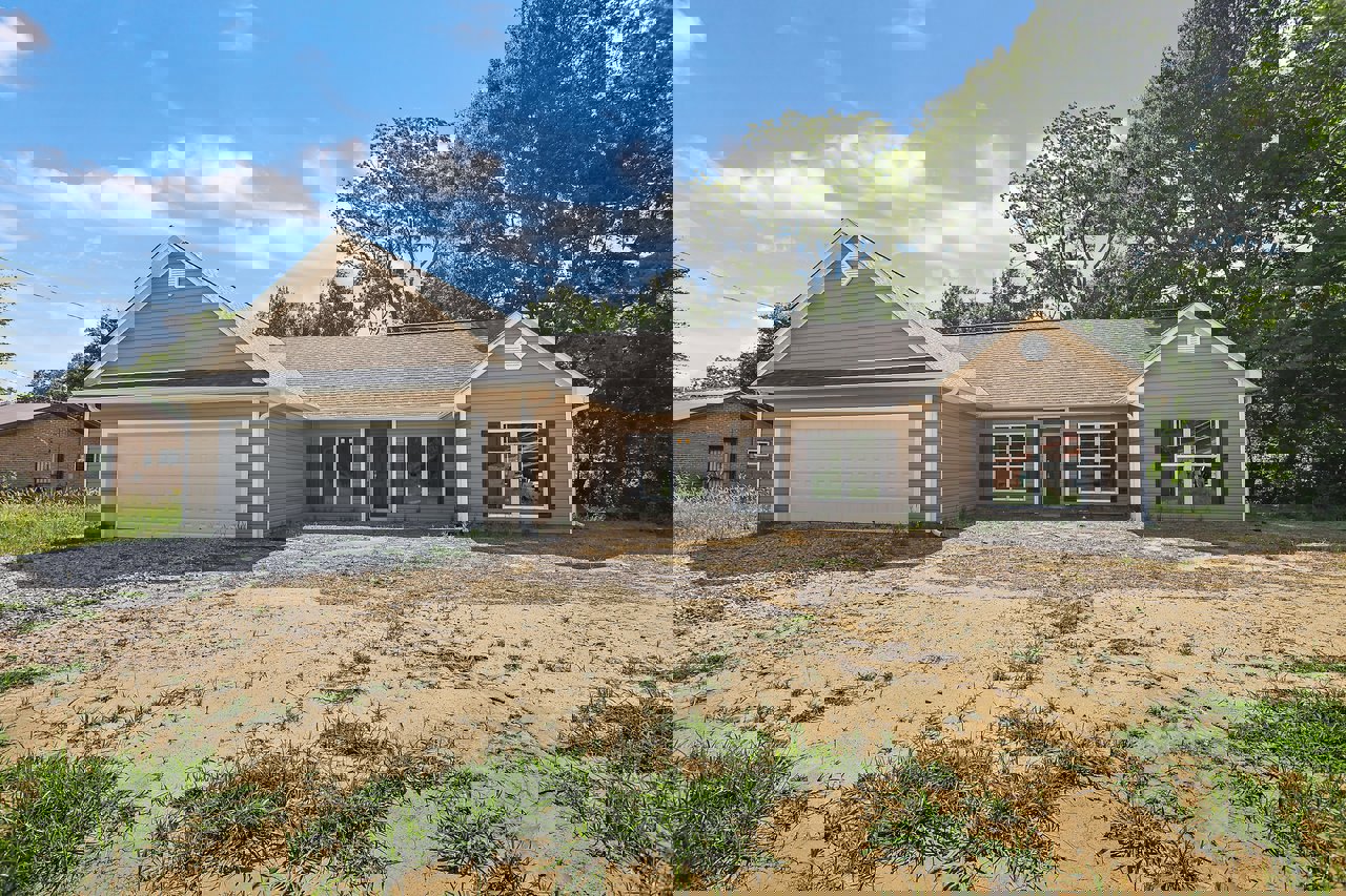 White garage door beneath brown roof, brick facade with front window and entry door, dirt patch and driveway leading to house, surrounded by trees under cloudy sky