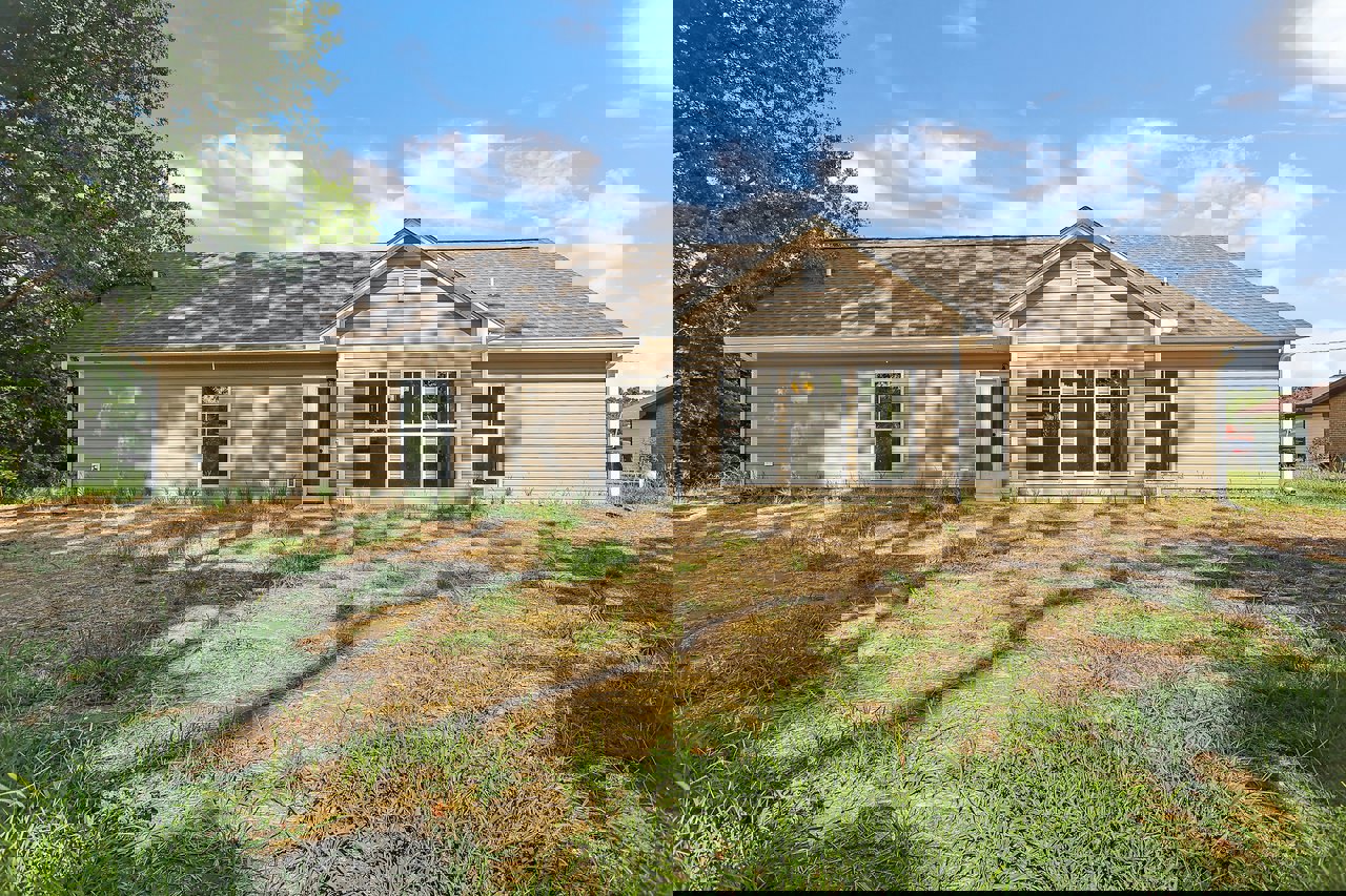 Two-story cottage-style home with white door and glass panel, row of windows, grassy yard bordered by trees, cloudy sky overhead, muddy patch near entrance