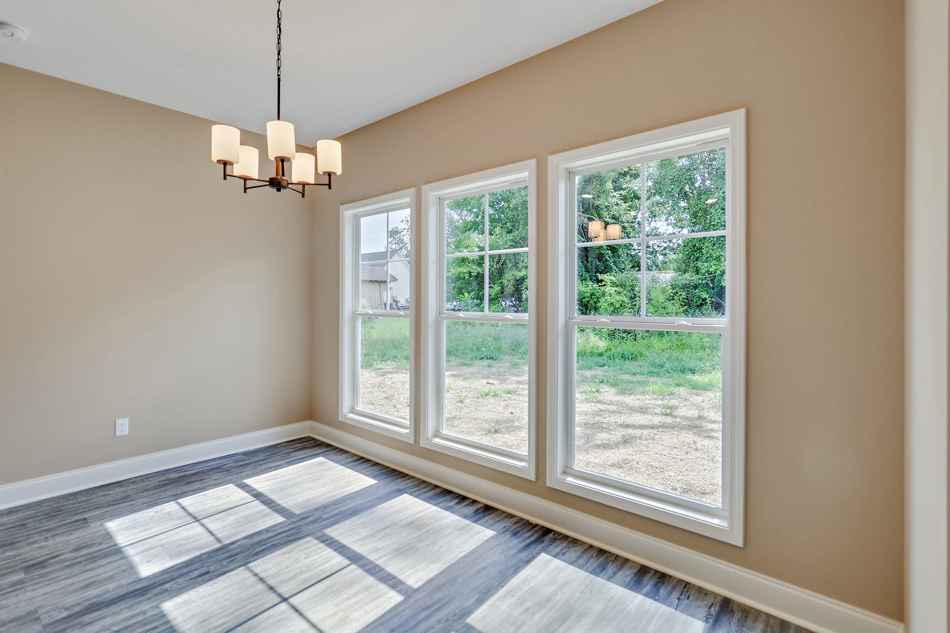 Spacious room with large windows, wood flooring, and a modern chandelier; sunlight streams in, highlighting neutral walls and a view of grass and trees outside.
