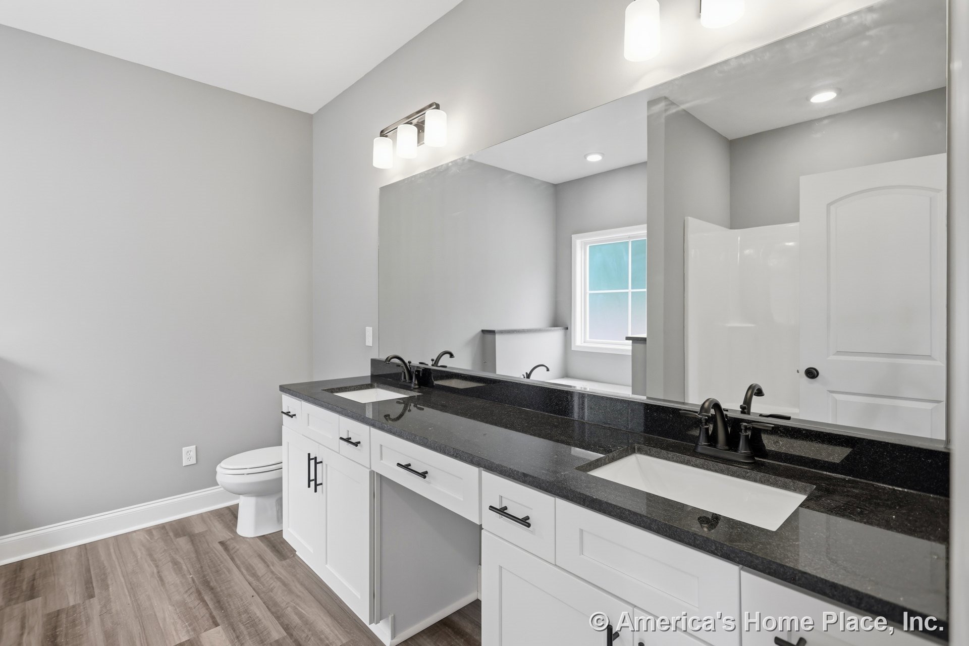 Double vanity with black stone countertops, white shaker cabinets, black faucets, large wall mirror, modern vanity lighting, wood-look flooring, shower stall with glass door