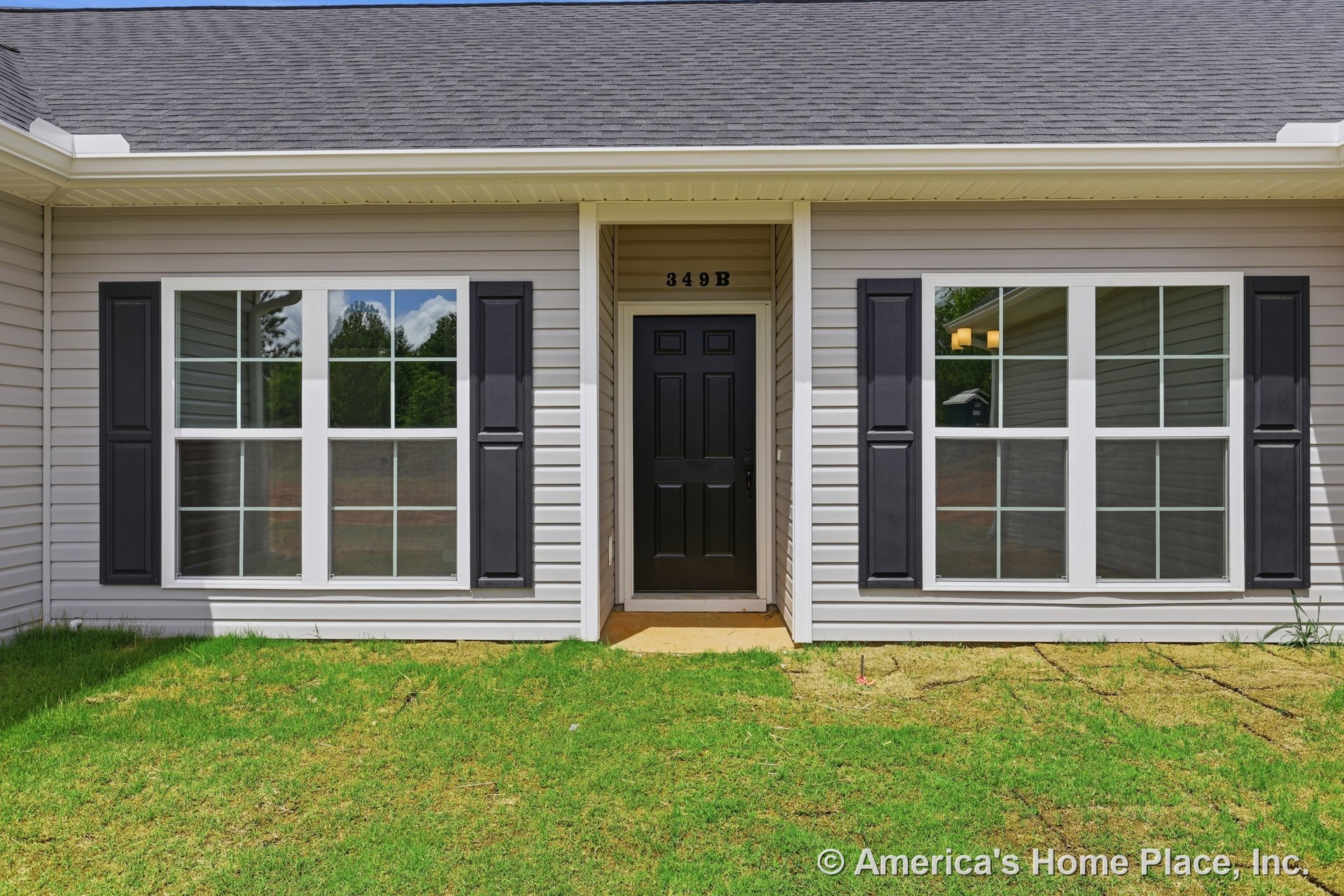 Black paneled front door with flanking double grid windows and matching black shutters, covered entry porch, light vinyl siding, asphalt shingle roof, and visible house number.