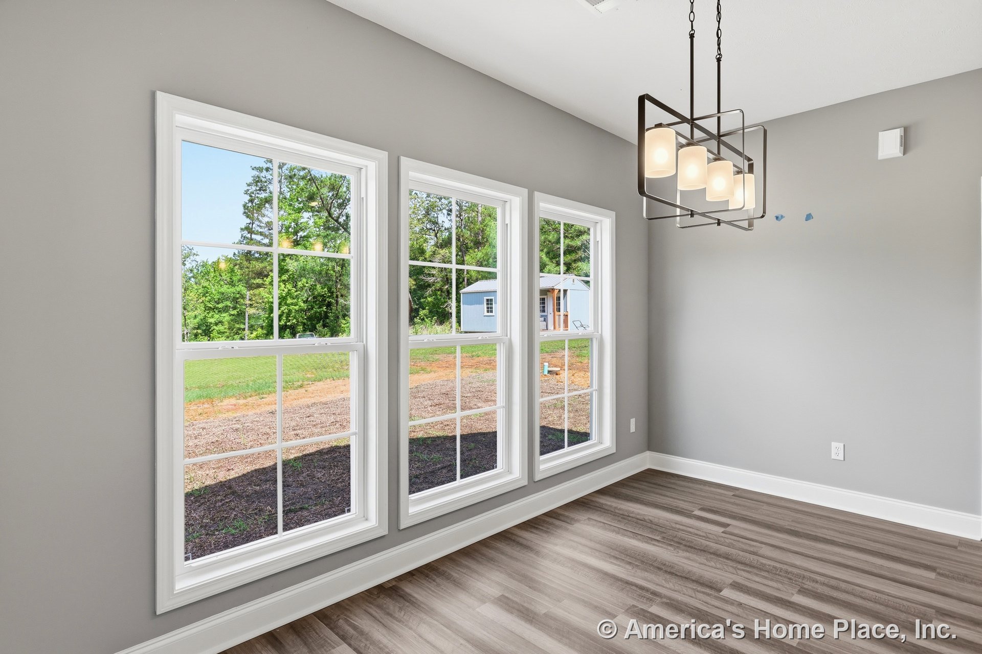 Dining area with three double-hung windows framed in white trim, gray painted walls, wood-look vinyl plank flooring, rectangular modern pendant chandelier, and white baseboards