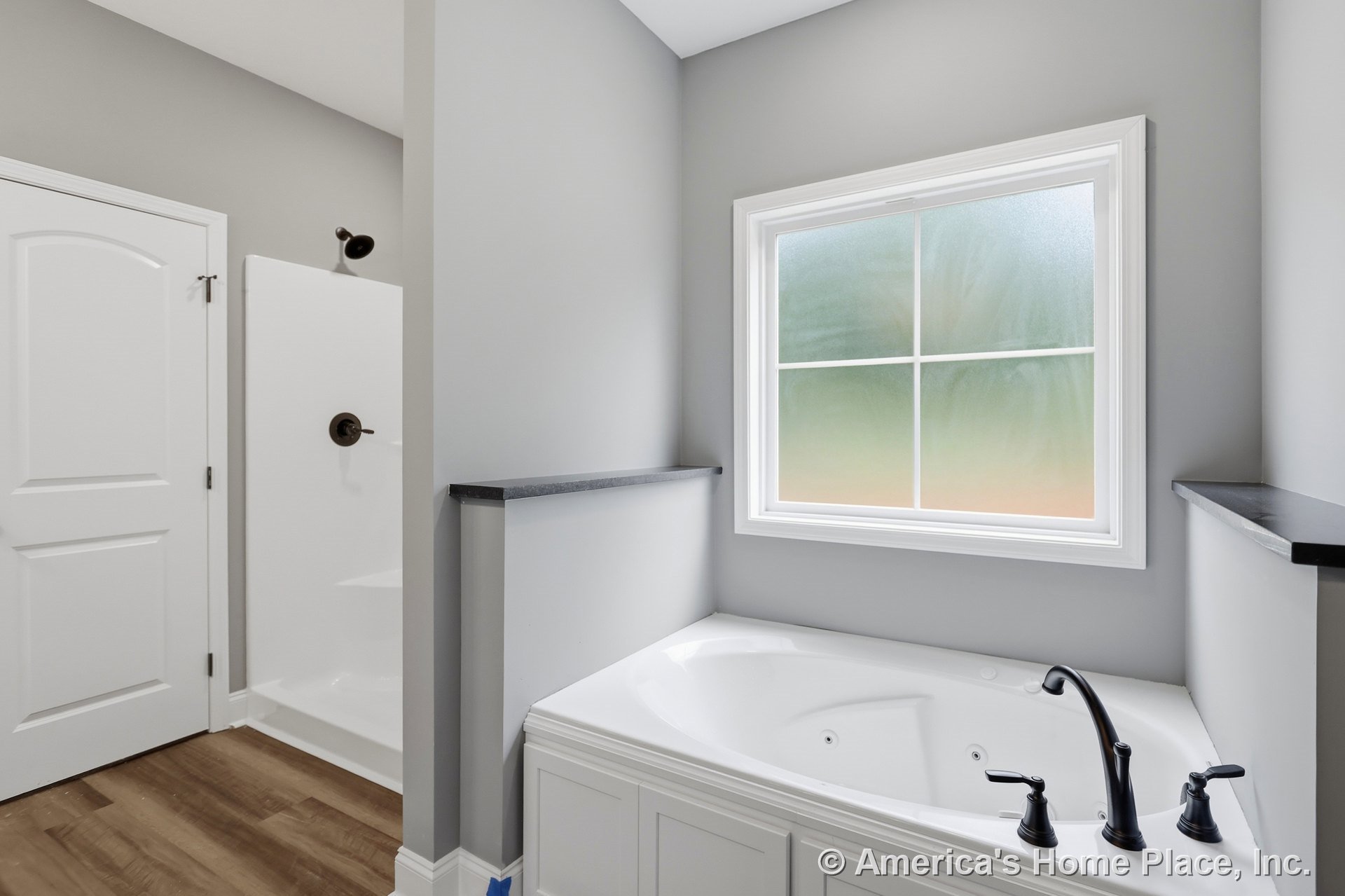Built-in white paneled soaking tub with black faucet beneath frosted window with white trim, adjacent walk-in shower enclosure, gray painted walls, wood-look flooring, and built-in