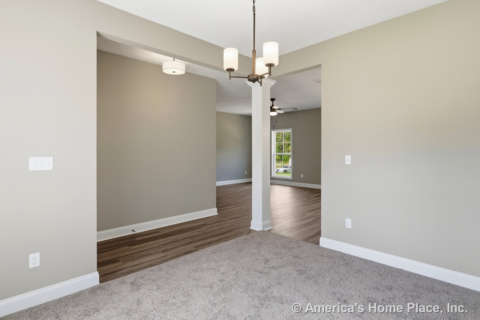 Open living area with neutral painted walls, central structural column, carpeted entry transitioning to wood-look flooring, modern chandelier lighting, large window framed in white
