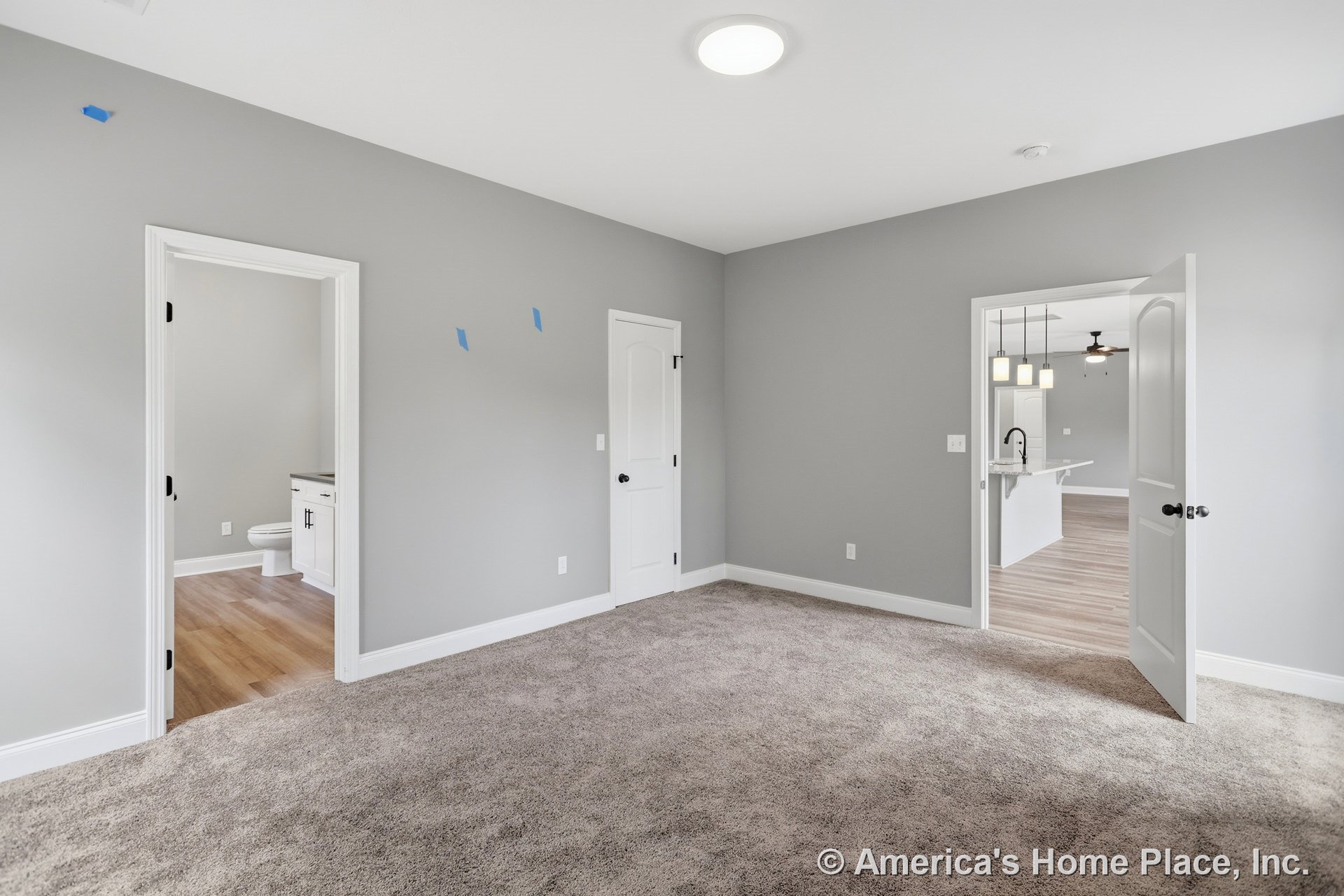 Carpeted bedroom with gray walls, white trim and baseboards, recessed ceiling light, white interior doors, visible bathroom vanity, and open doorway leading to kitchen area.