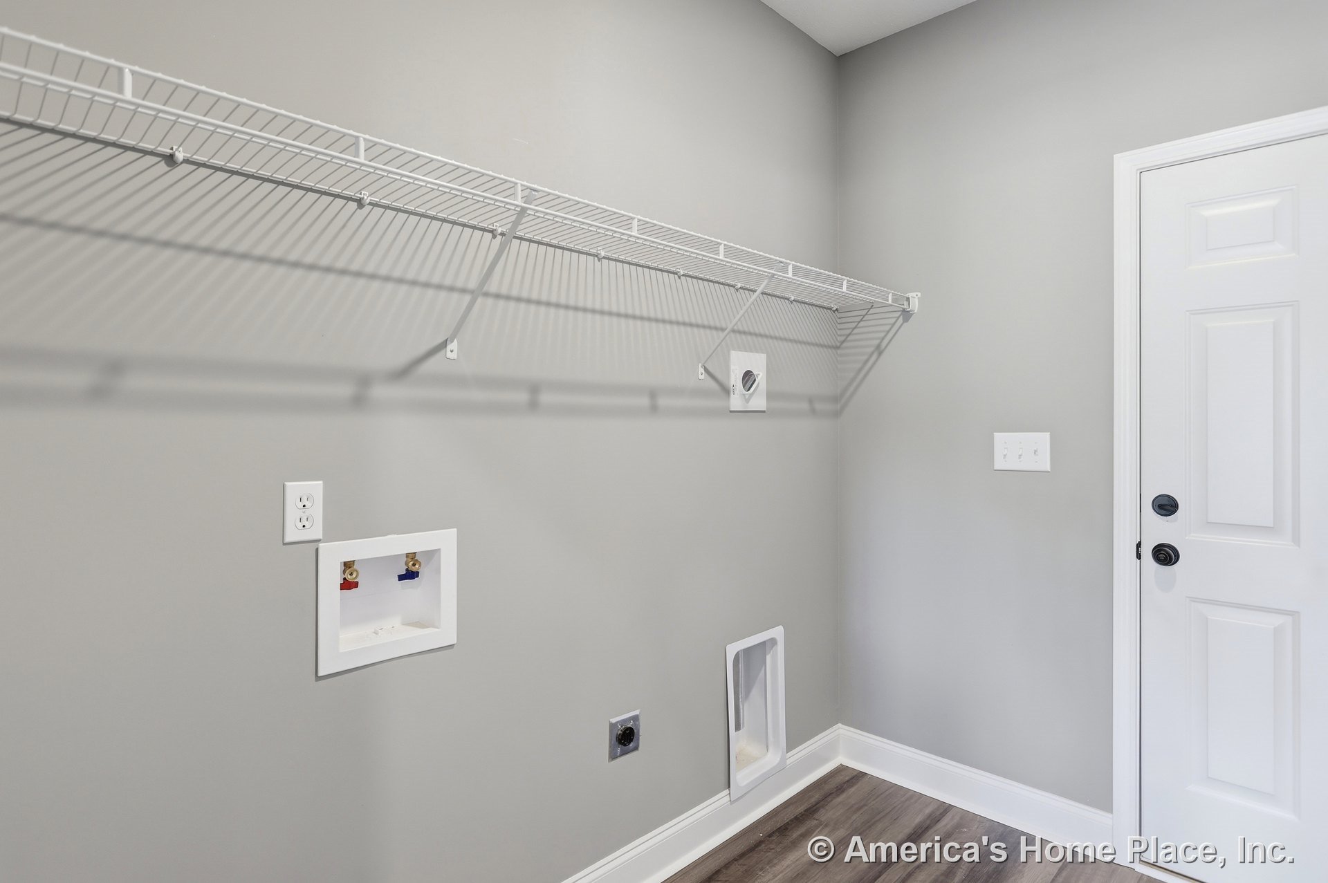 Laundry room with wall-mounted wire shelf above washer and dryer hookups, gray painted walls, white baseboard trim, wood-look vinyl flooring, utility outlet, wall vent, and white