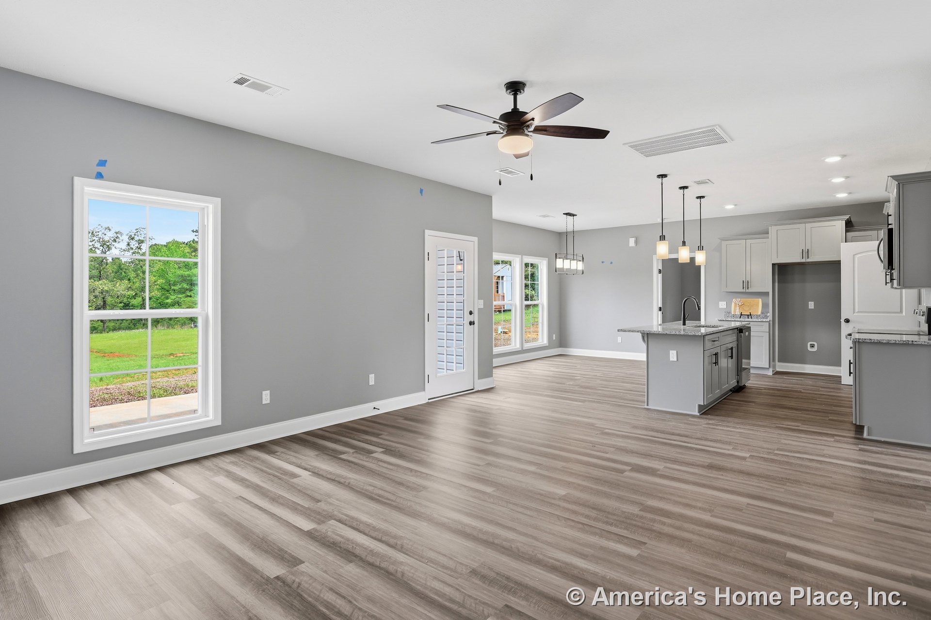 Open-concept living area with light wood-look flooring, gray shaker kitchen cabinets, granite countertops, kitchen island with pendant lights, large windows with white trim