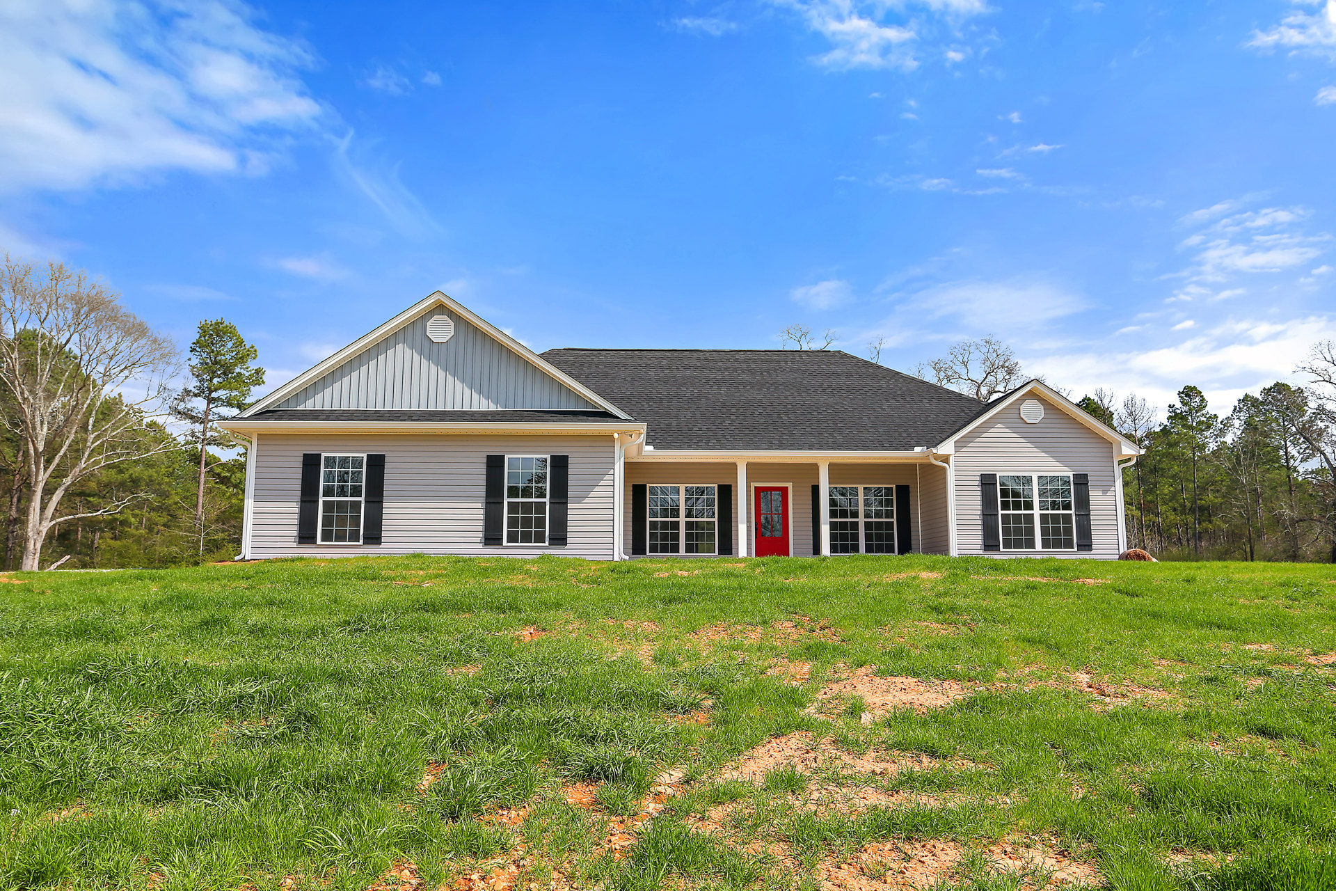 White cottage-style home with black shuttered windows, red front door featuring a glass pane, manicured green lawn, leafless tree in foreground, partly cloudy sky overhead