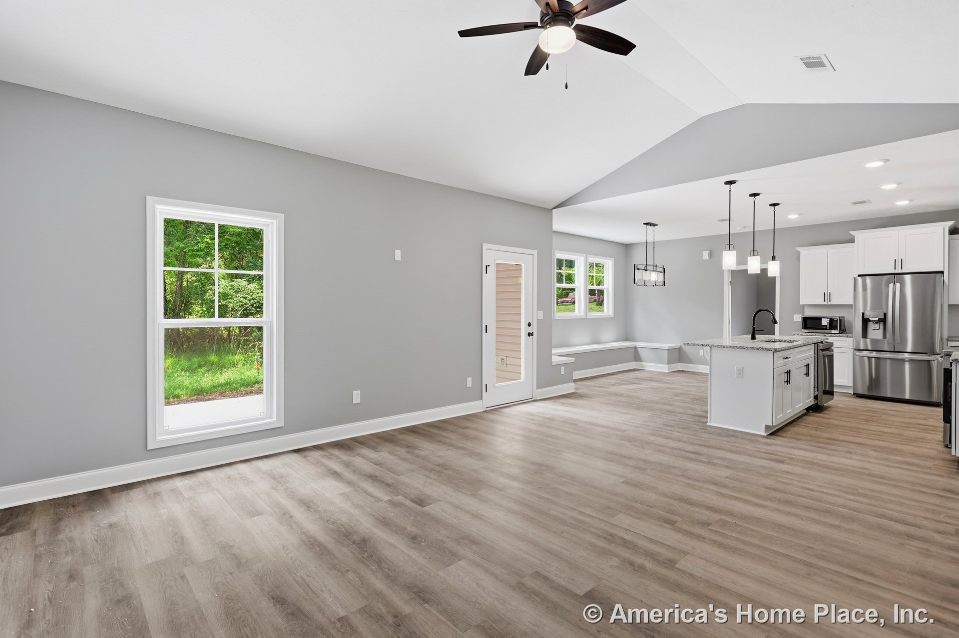 Open-concept living area with light wood plank flooring, vaulted ceiling and ceiling fan, large window with white trim, modern kitchen featuring white cabinets, granite