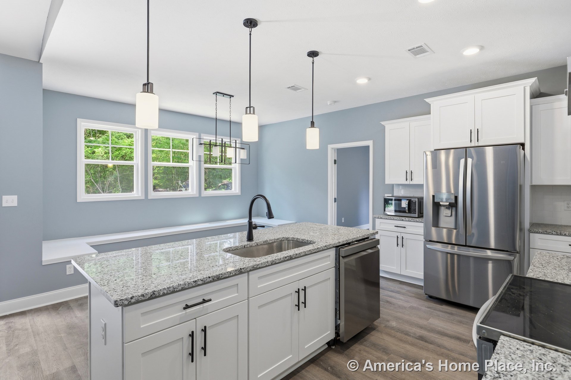White shaker cabinets and granite countertops surround a spacious kitchen island with sink, pendant lighting fixtures overhead, stainless steel refrigerator and built-in microwave