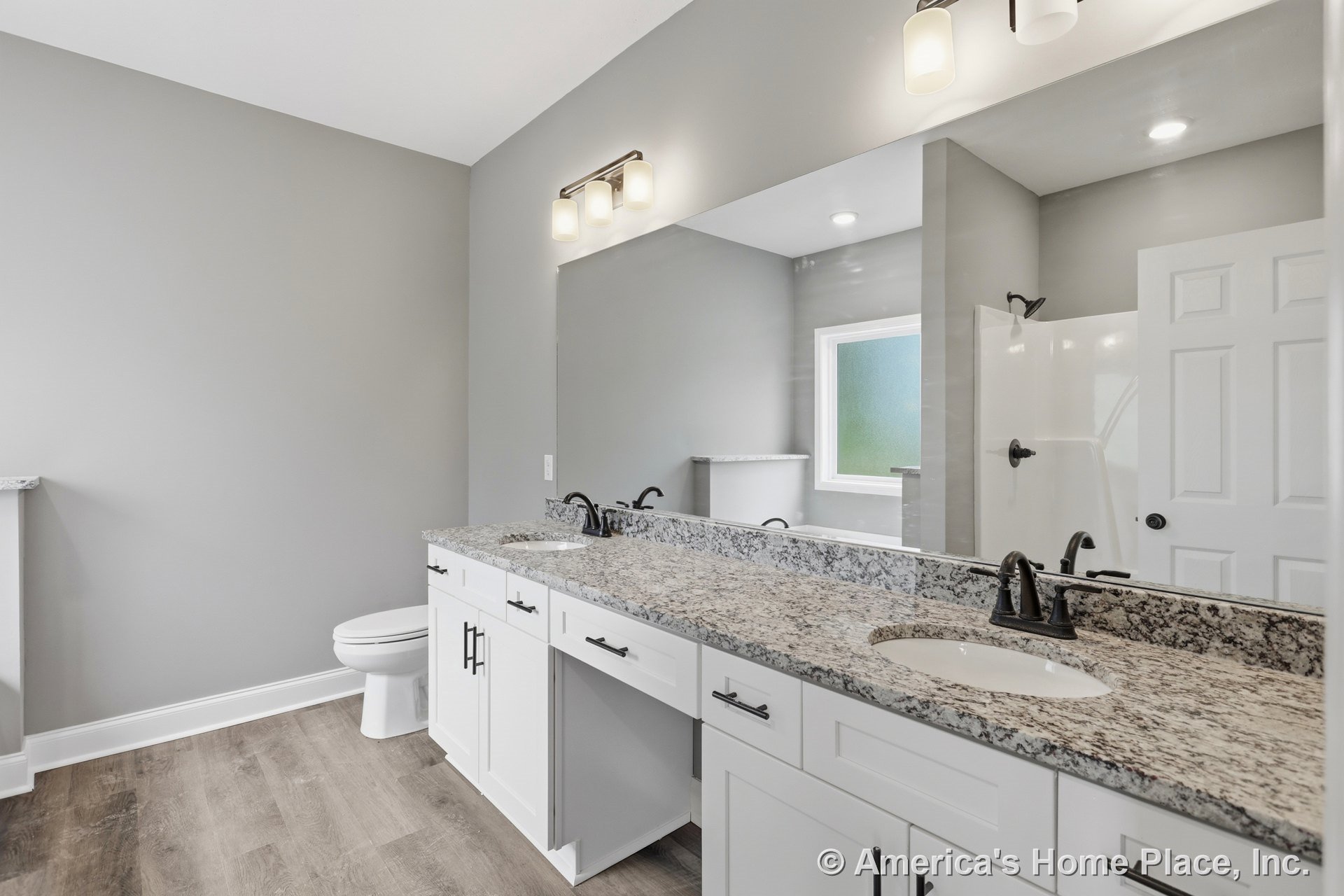 Double vanity with granite countertop, white shaker cabinets, double sinks, large wall mirror, built-in overhead lighting, walk-in shower with frosted window, gray walls, wood-look