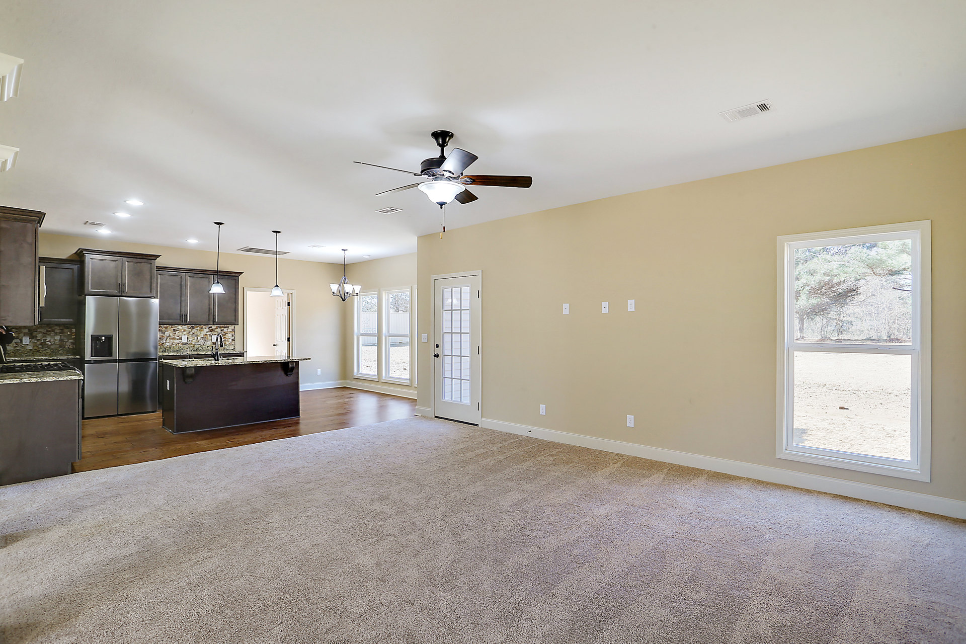 Open concept living room with ceiling fan and light fixture, adjoining kitchen featuring black cabinetry, granite countertops, stainless steel refrigerator with glass door, and