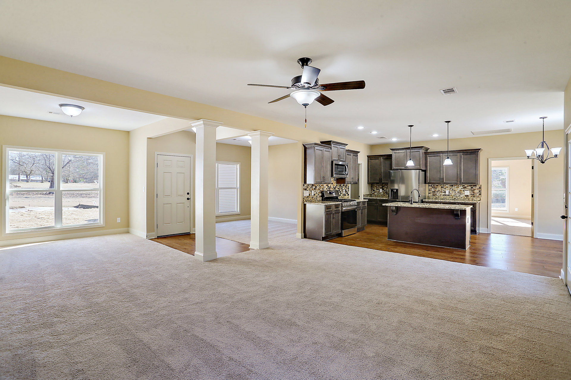 Open-concept living room with granite kitchen countertops, ceiling fan with light fixture, white door with black knobs, large window overlooking field and trees, neutral walls and