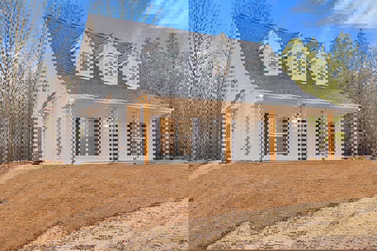 White siding house with black shingle roof, covered front porch, leafless trees in background, brown grass and dirt yard, windows with closed blinds, large gray HVAC unit near