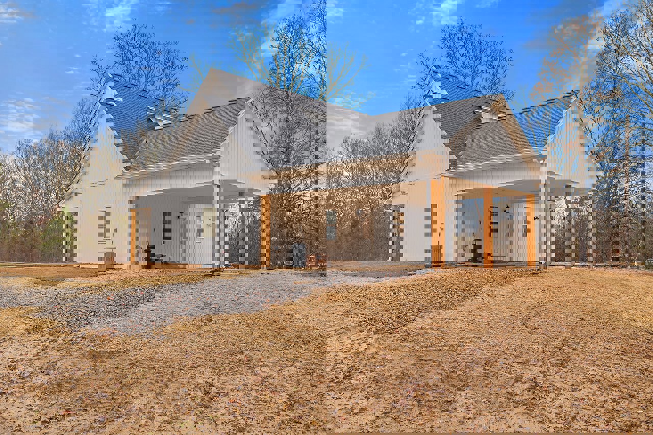 White house with black roof, large covered porch, attached garage, expansive driveway, leafless tree, dirt and gravel area, cloudy sky