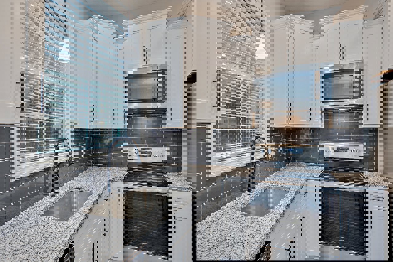 White kitchen cabinets, stainless steel sink with black faucet, glass door stove, built-in microwave, light fixture on ceiling, stone countertop.