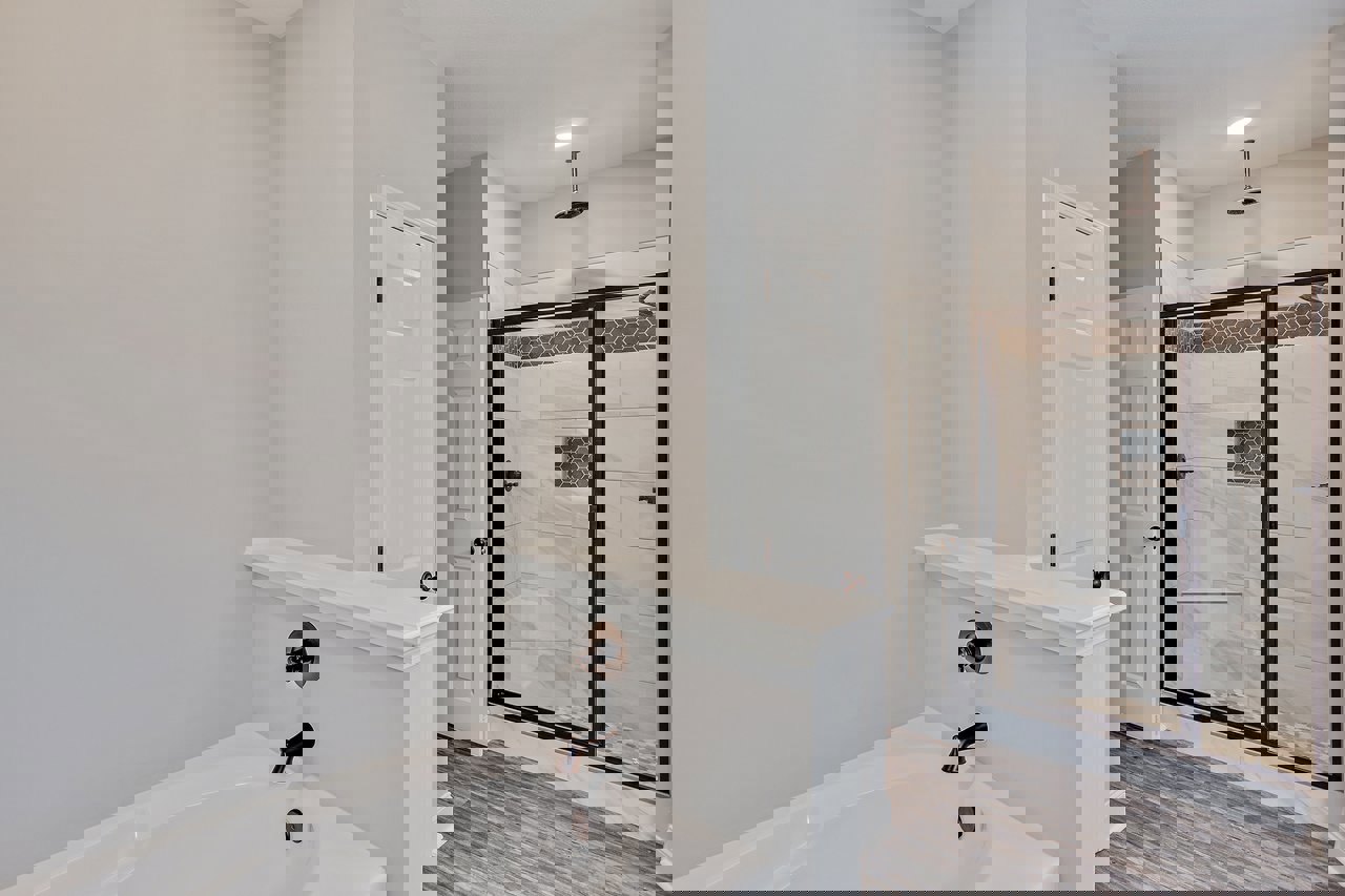 White bathtub with chrome faucet next to glass-enclosed shower, gray tile walls, and black soap dispenser on countertop