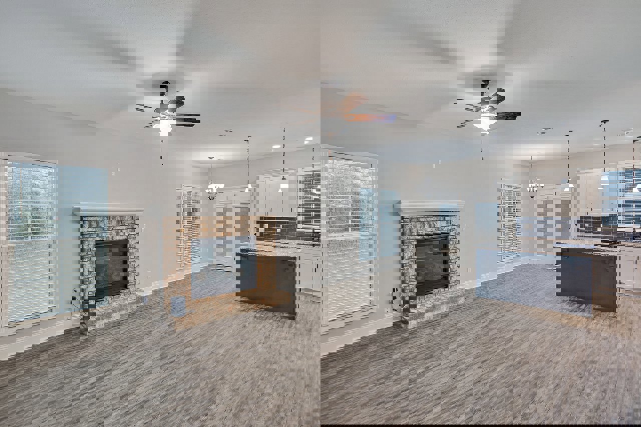 Open concept living room with brick fireplace featuring black glass, marble countertop in adjacent kitchen, window with blinds, ceiling fan with light fixture, hardwood flooring