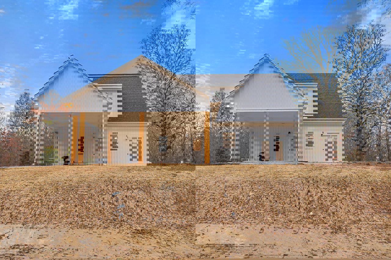 White custom home under construction with exposed dirt lot, leafless tree nearby, visible roof, windows, and white door with glass insert.