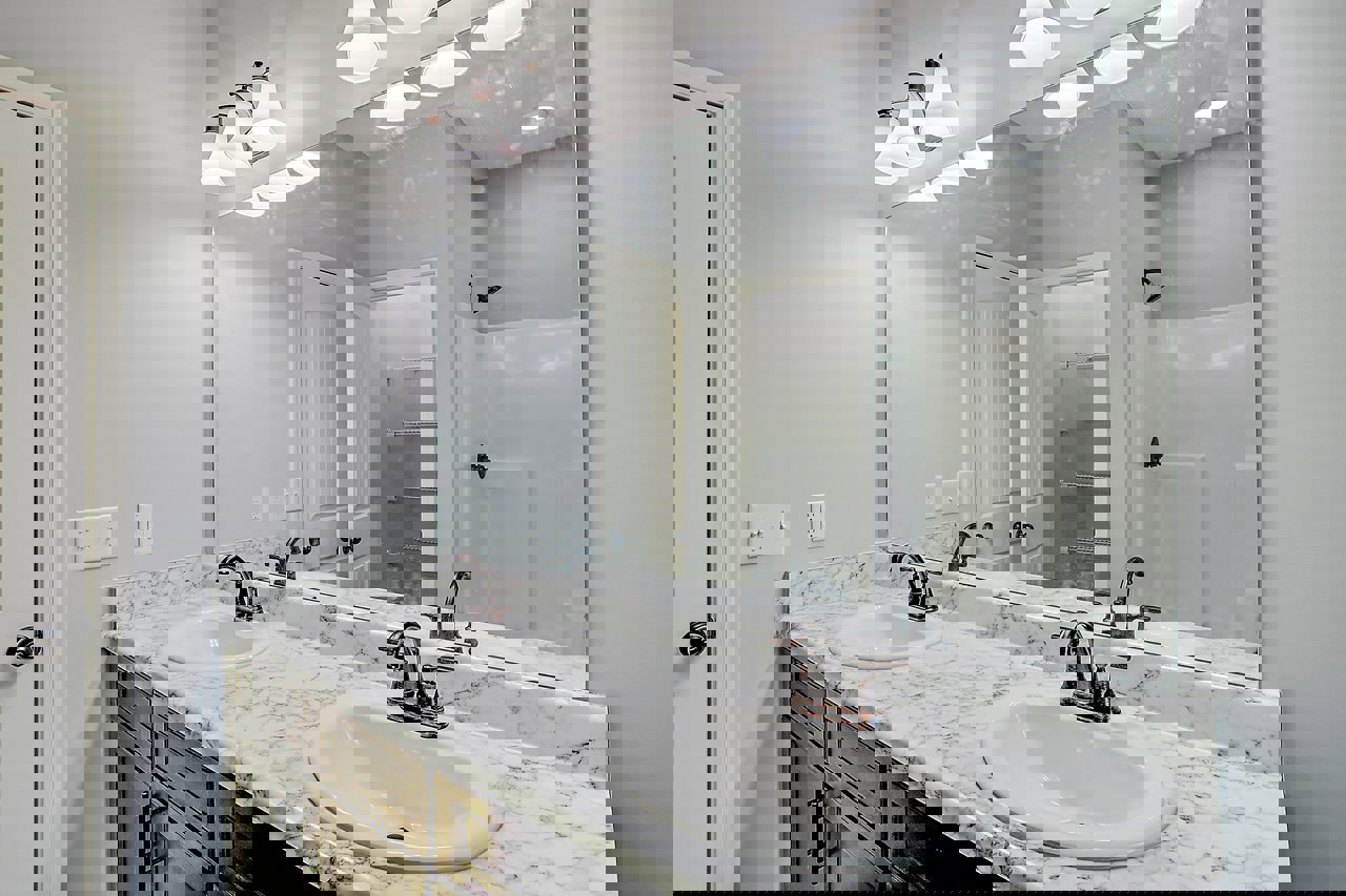 Bathroom with marble countertop, dual sinks, chrome faucets, large wall-mounted mirror, white walls, and modern fixtures