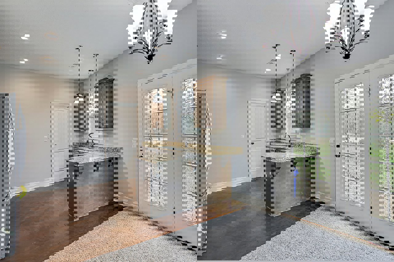 Granite kitchen countertops, brown cabinetry, tile flooring, stainless refrigerator beside a person, glass-paned door leading outside, close-up chandelier, white lamp on cabinet