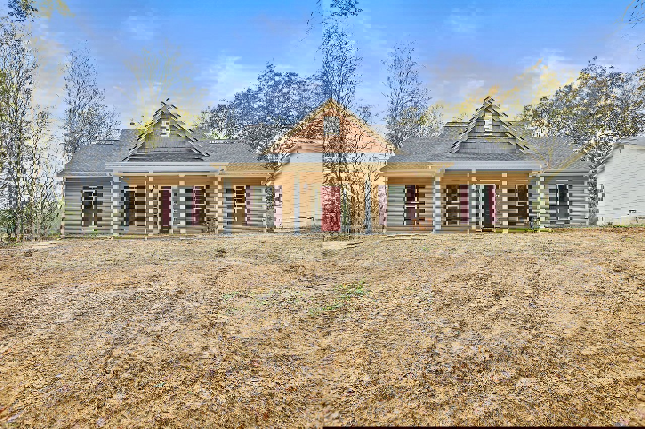 White house with red door featuring glass panes, large grassy yard, white-framed windows, gray roof with vent, surrounded by trees and plants under partly cloudy sky