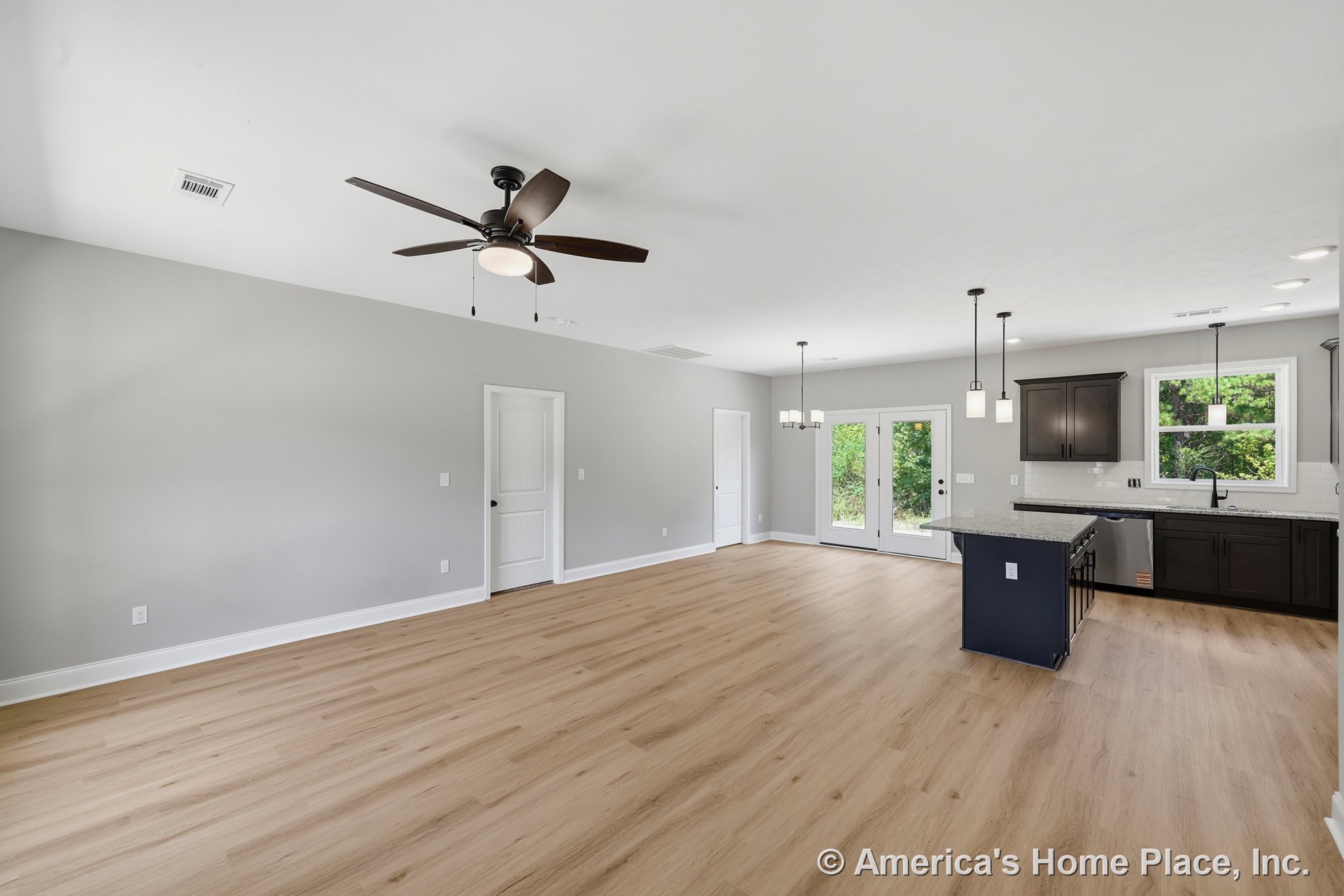 Open-concept living area with light wood plank flooring, dark kitchen cabinets, granite island countertop, pendant and recessed lighting, ceiling fan, double glass patio doors, and