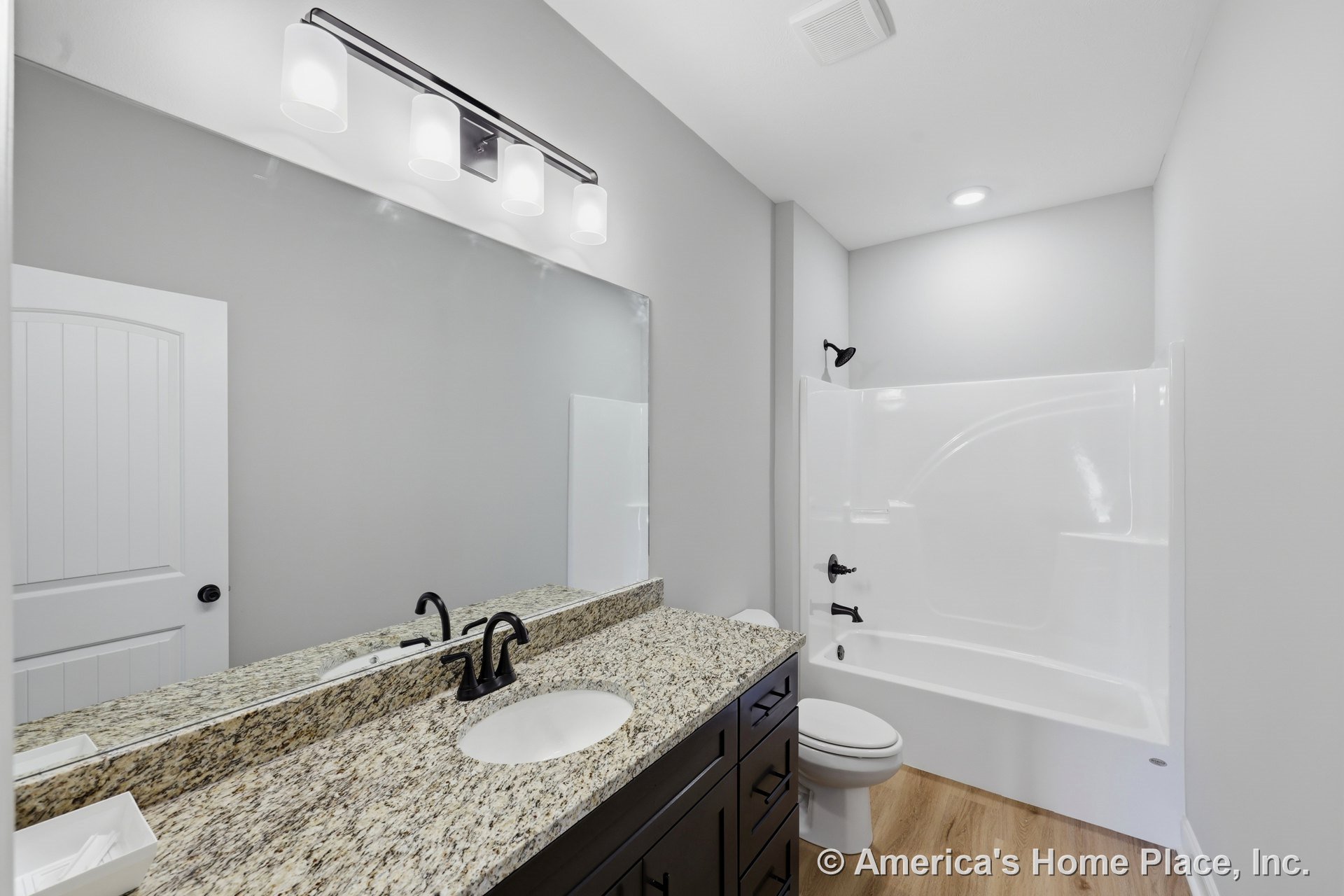 Granite vanity with dark wood cabinets, matte black fixtures, fiberglass tub-shower combo, wood-look flooring, large wall mirror, recessed lighting, wall sconce, and white walls.