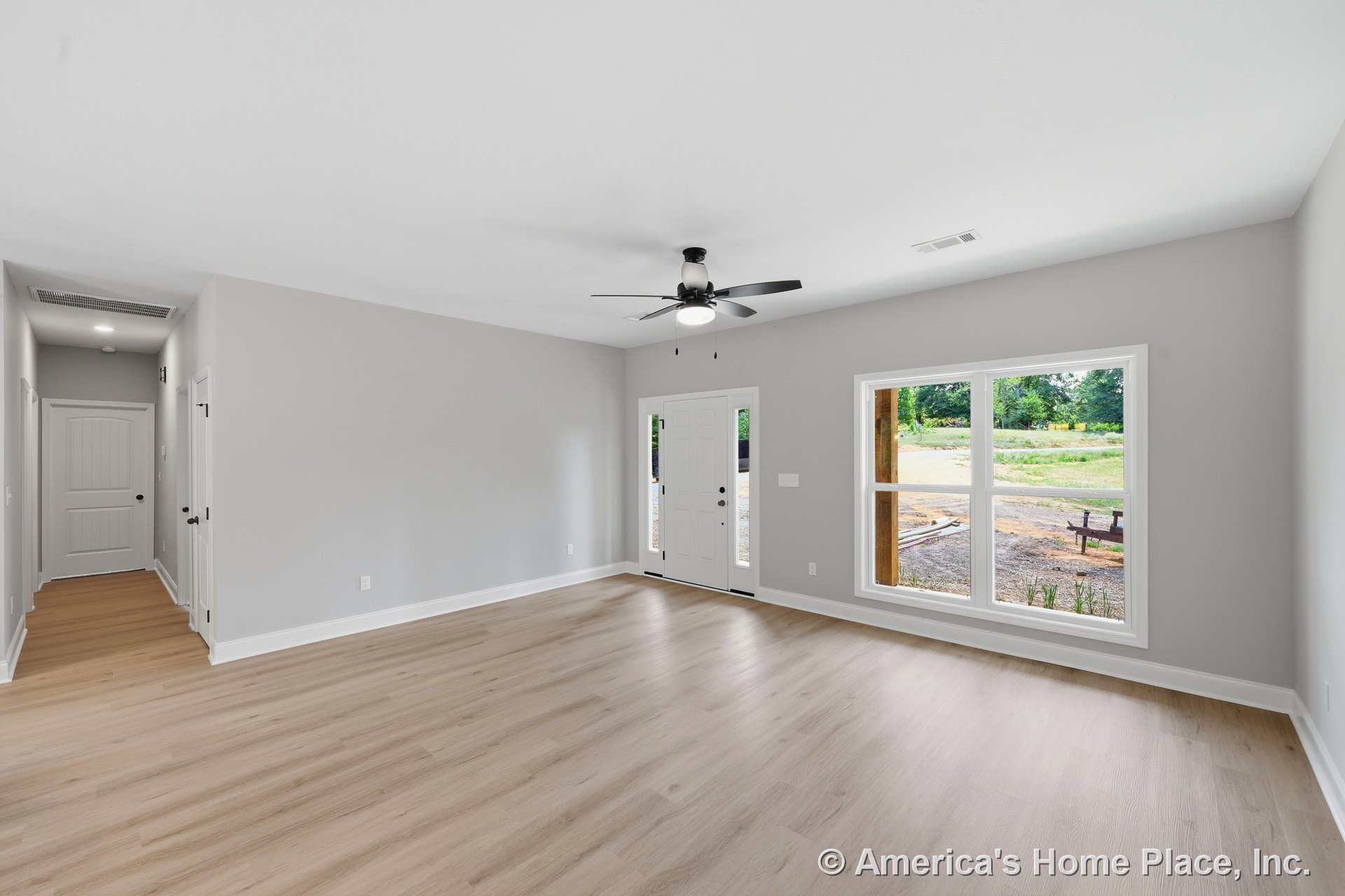 Light wood plank flooring, large double-pane picture window with white trim, gray painted walls, ceiling fan with built-in lighting, entry door featuring a glass insert, and modern