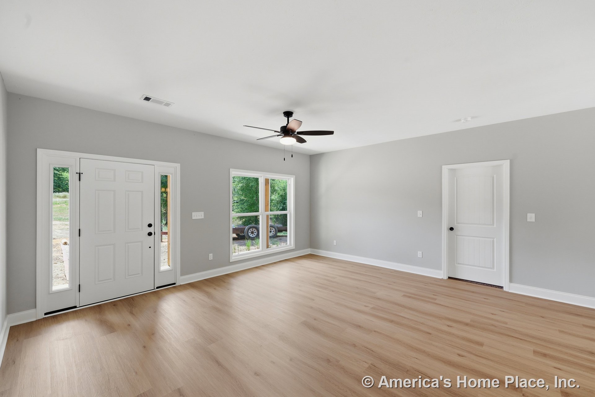 Living room with light wood plank flooring, gray walls, large front windows, white paneled front door, side entry door, baseboard trim, and ceiling fan with integrated lighting.