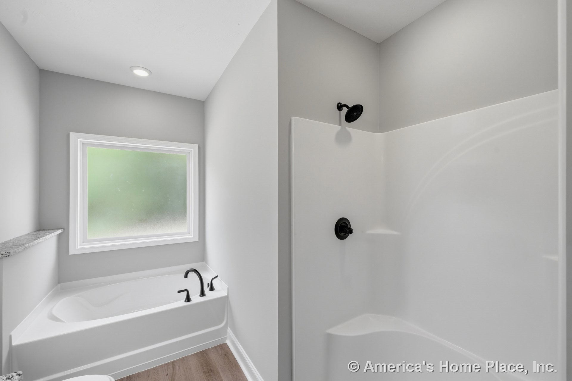 Built-in soaking tub with matte black faucet beside a separate glass shower enclosure featuring matching black fixtures, frosted window with white trim, recessed ceiling light