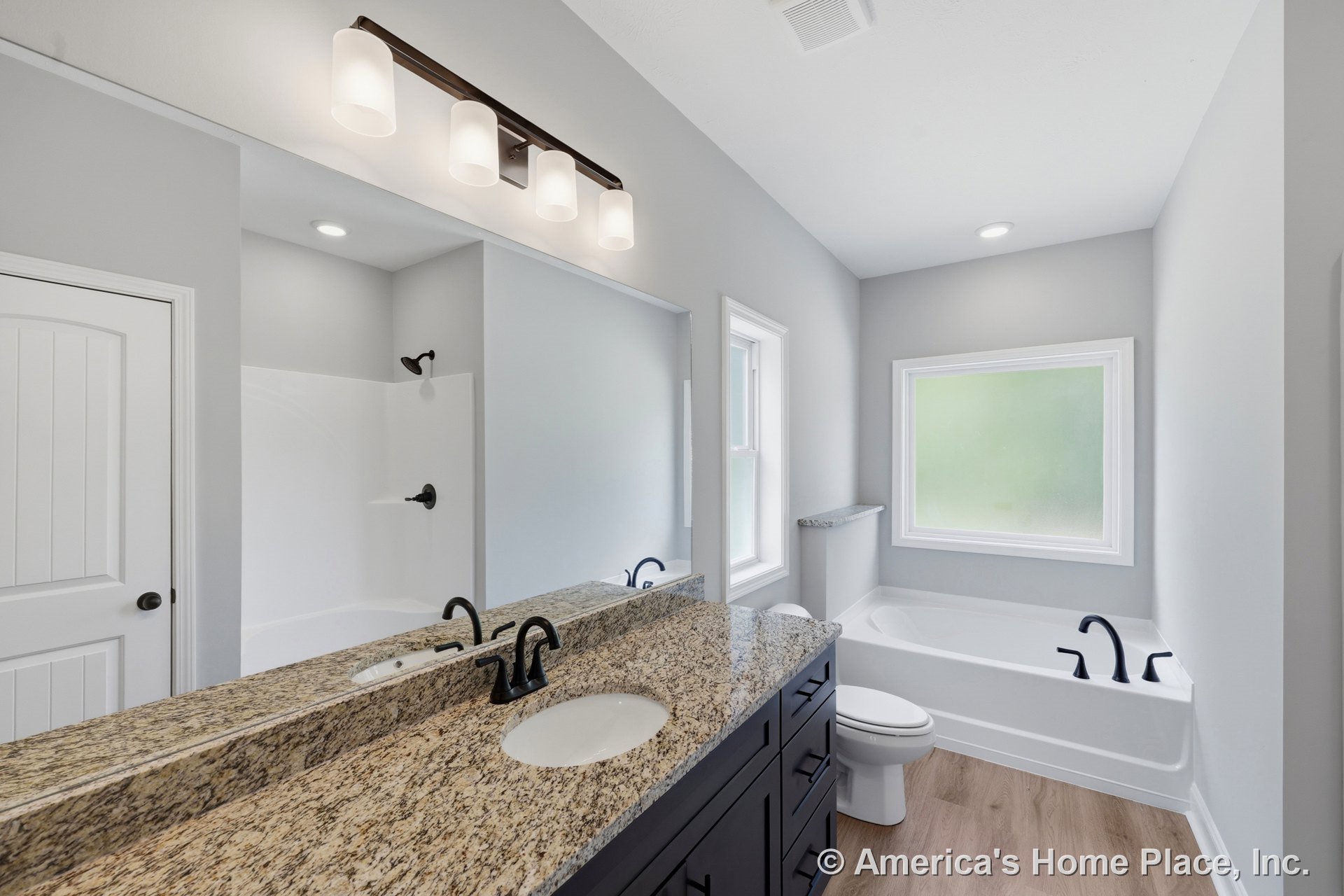 Granite countertop vanity with integrated sink and black fixtures, built-in bathtub, large frosted window, wood-look flooring, white paneled door, modern overhead lighting, neutral