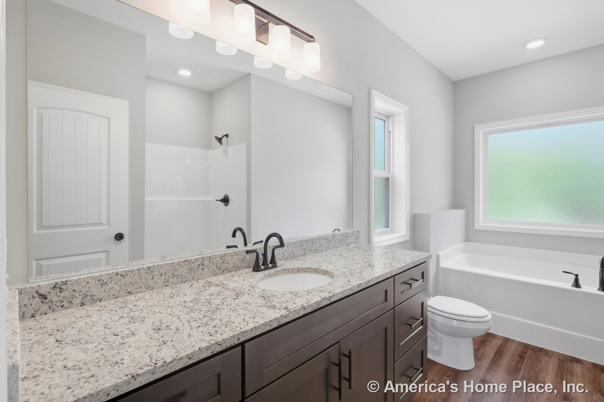 Granite countertop vanity with dark wood cabinetry, single sink, matte black faucet, large mirror, soaking tub with matching matte black fixtures, frosted glass window, wood-look