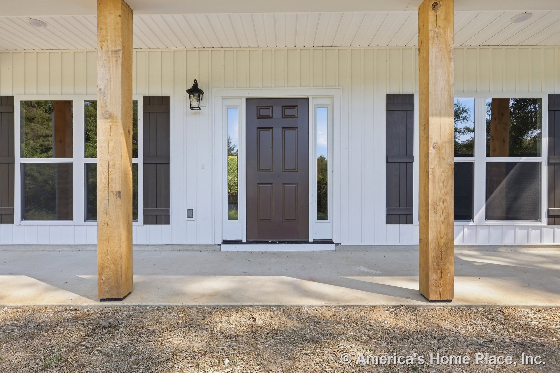Wood porch columns frame a brown paneled front door with sidelights, vertical white siding, double windows with shutters, concrete porch floor, and exterior wall lantern.