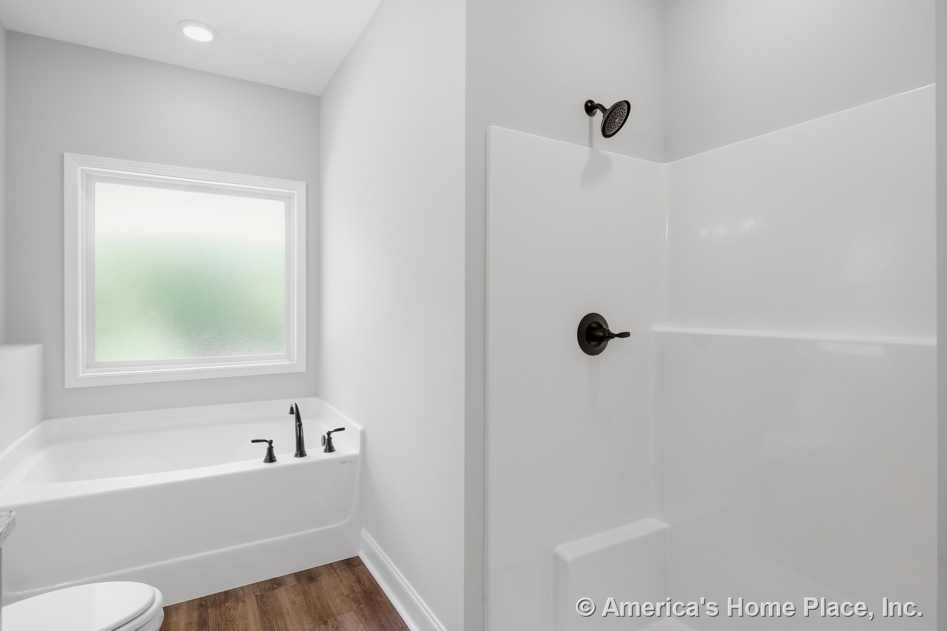 Built-in white acrylic bathtub beside a separate shower enclosure with oil-rubbed bronze fixtures, frosted window, recessed ceiling light, wood-look vinyl flooring, and white trim.