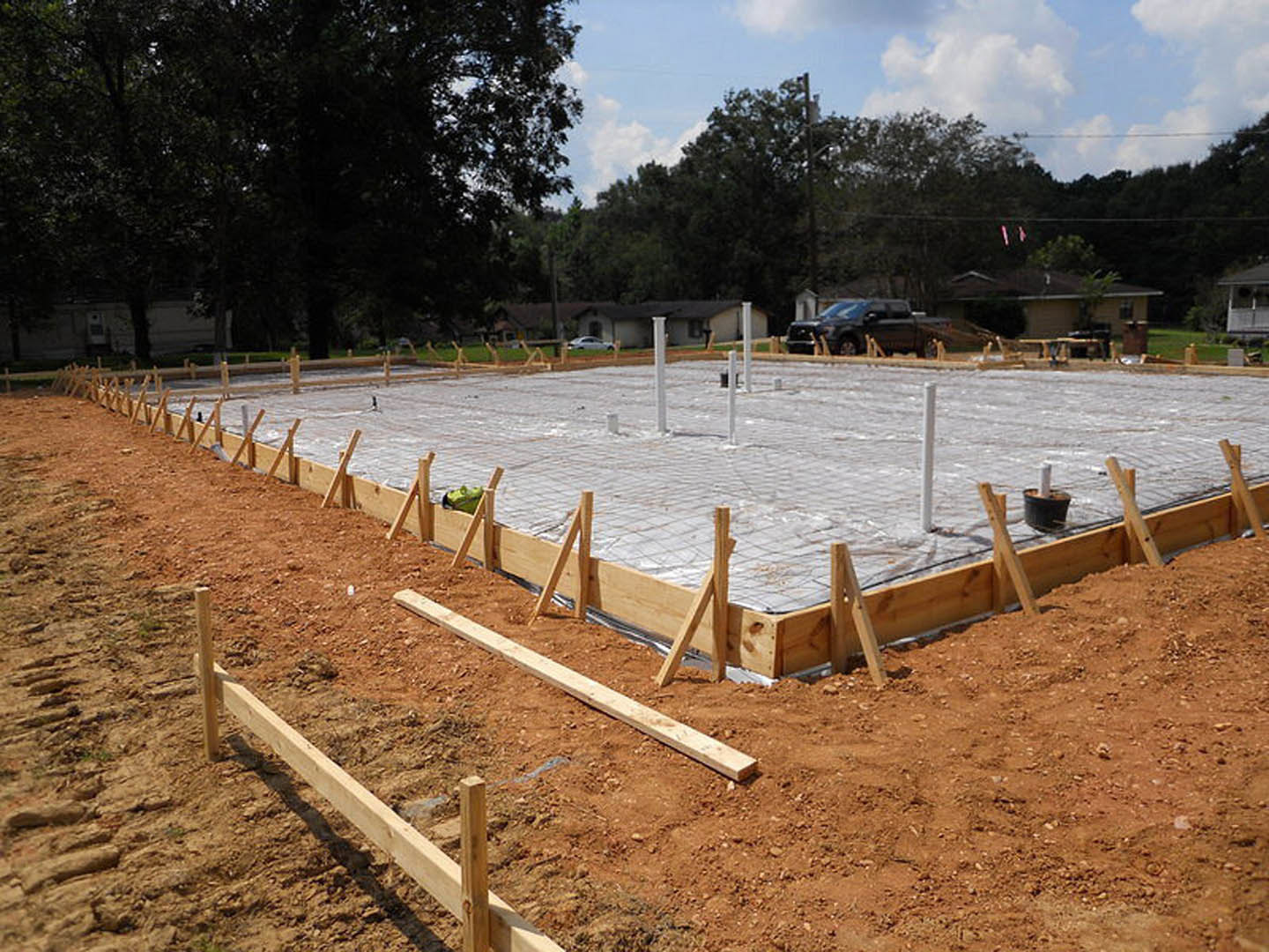 Wood and concrete foundation under construction with wooden beams and poles, black truck in background, trees and cloudy sky surrounding site